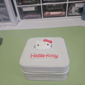 May include: A white, square jewelry box with a zipper closure. The top features a Hello Kitty design with a red bow and the words "Hello Kitty" in pink. The box is sitting on a green surface.