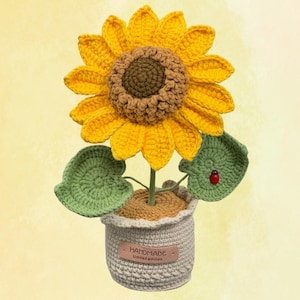 Pode incluir: Um girassol de crochê feito à mão em um vaso pequeno. O girassol tem pétalas amarelas brilhantes e um centro marrom. Folhas verdes com uma joaninha vermelha acentuam a flor. O vaso é cinza claro com uma etiqueta "HANDMADE Limited edition".