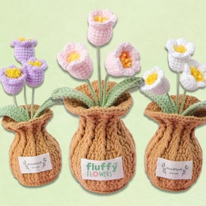 Könnte beinhalten: Drei gehäkelte Blumenarrangements in beigen Töpfen. Die Blumen sind in Lila-, Rosa- und Weißtönen mit gelben Zentren und grünen Blättern gehalten. Die Töpfe tragen Etiketten mit der Aufschrift "Handmade with love" und "Fluffy Flowers".