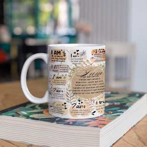 May include: White ceramic mug with a motivational quote that reads "I am" and lists positive affirmations. The mug is decorated with floral patterns and hearts.