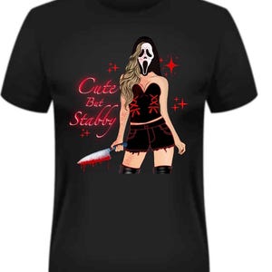 May include: Black t-shirt featuring a graphic of a woman in a Scream mask holding a bloody knife. The design includes the text "Cute But Stabby" in red, with red star accents. The woman is wearing a black corset, shorts, and boots.