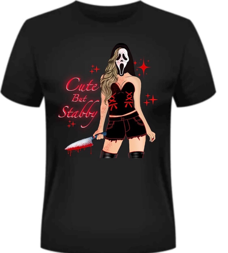 May include: Black t-shirt featuring a graphic of a woman in a Scream mask holding a bloody knife. The design includes the text "Cute But Stabby" in red, with red star accents. The woman is wearing a black corset, shorts, and boots.