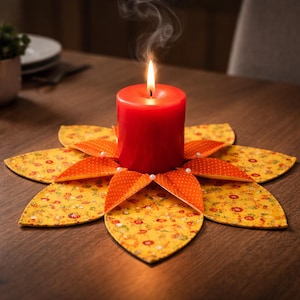 May include: A lit red candle sits atop a decorative, flower-shaped candle holder. The holder is made of yellow fabric with a floral pattern and orange polka-dot accents. Small white beads are placed around the candle.
