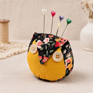 May include: A fabric owl-shaped pincushion featuring a black floral print top and a solid yellow bottom. Two beige buttons serve as eyes. Four straight pins with heart-shaped heads in white, red, purple, and green are inserted into the top.