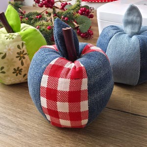 May include: Three fabric apple-shaped ornaments. One is light green with white daisy patterns, one is blue denim with a red and white checkered panel, and one is solid blue denim. Each features a brown cinnamon stick as a stem.