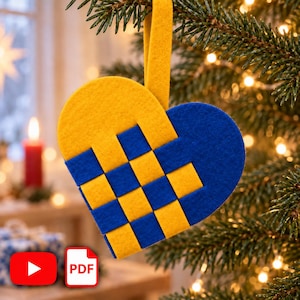 Op de afbeelding: Een hartvormig ornament gemaakt van geweven gele en blauwe viltstroken. Het ontwerp heeft een rasterpatroon met een bijpassende gele viltlus aan de bovenkant om op te hangen. Het ornament heeft een levendig, tweekleurig kleurenschema.