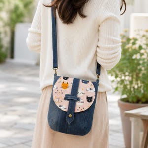 May include: A denim crossbody bag with a flap featuring a cat pattern. The bag has a blue denim body, a pink flap with cat faces in orange, black, and white, and a long, adjustable strap.