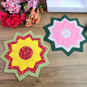 Può includere: Due sottopentola fatti a mano a forma di stella con disegni floreali. Uno presenta un centro giallo con dettagli rossi e verdi, l'altro un centro rosa con dettagli bianchi e verdi. I sottopentola sono su una superficie di legno.