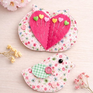 May include: Two handmade pin cushions. One is heart-shaped, made of pink felt with colourful heart-shaped pins. The other is bird-shaped, with a floral pattern and a green gingham wing. Both have button details.