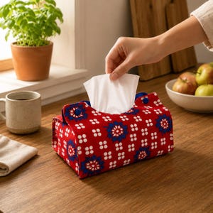 May include: A rectangular fabric tissue box cover featuring a vibrant red background with a repeating pattern of white dots and blue floral motifs. The cover has blue snap buttons on the top flaps and holds a stack of white facial tissues.