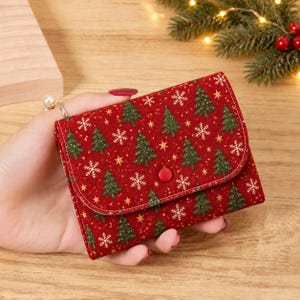 May include: A red wallet featuring a repeating pattern of green Christmas trees, white snowflakes, and small gold stars. The wallet has a curved flap closure secured with a single red button snap, and it is constructed with visible white stitching.
