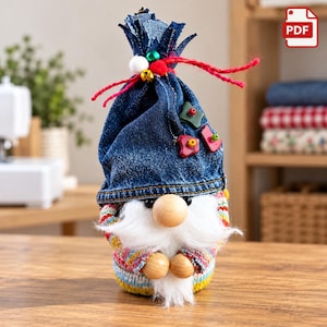 Könnte beinhalten: Eine Gnomenfigur mit einer dunkelblauen Jeansmütze, die oben mit einer roten Schnur zusammengebunden und mit kleinen bunten Knöpfen verziert ist. Der Gnom hat eine runde Holznase, einen weißen Kunstfellbart und einen Körper aus bunt gestreiftem Strick.