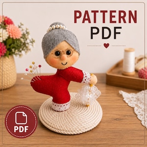 Op de afbeelding: Een viltpop van een ouder persoon met grijs haar, een rood shirt met lange mouwen en witte kanten rand, een ronde bril en parels. De pop houdt een wit kanten parasolletje vast en dient als speldenkussen met gekleurde spelden. Tekst: PATTERN PDF.