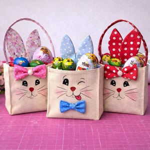 Op de afbeelding: Drie paasmandjes met konijnengezichten, oren en handvatten. Elk mandje is gevuld met versierde paaseieren. De mandjes hebben roze, blauwe en rode strikken. De konijnenoren zijn voorzien van stippen en bloemmotieven.