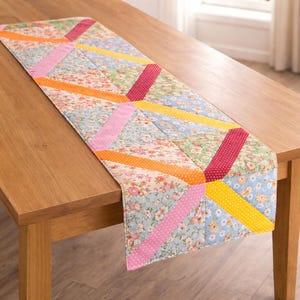 May include: A colorful quilted table runner with a floral pattern and diagonal stripes in pink, orange, yellow, and red. The runner is placed on a wooden table, showcasing its patchwork design.