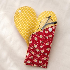 May include: A heart-shaped fabric glasses case featuring a red exterior with a white daisy and polka dot pattern, and a yellow interior with white polka dots. The case is secured with a white snap button and holds a pair of black-framed glasses.