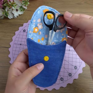 Puede incluir: Un estuche de tela para tijeras con interior azul claro con estampado floral naranja y blanco y exterior azul sólido. El estuche incluye un cierre de presión amarillo y contiene un par de tijeras de costura pequeñas de metal con mangos negros.