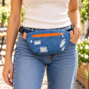 Pode incluir: Uma pochete de ganga azul com textura desgastada e desfiada, um bolso frontal com fecho de correr laranja e painéis laterais decorados com um padrão floral branco sobre fundo laranja. O saco inclui uma alça preta ajustável com fivela.