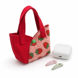 Può includere: Una piccola borsa in tessuto con un lato rosso e un lato rosa con motivo a fragole, chiusa da un bottone a pressione verde. Accanto alla borsa si trovano una custodia di ricarica bianca per auricolari wireless, una molletta rosa e una verde chiaro.