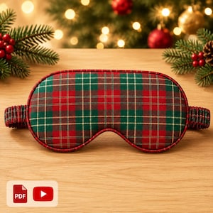 May include: A sleep mask featuring a classic red and green plaid pattern with thin gold accent lines. The mask has a rounded, contoured shape with a matching fabric-covered elastic strap on each side to secure it in place during use.