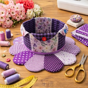 May include: A handmade purple and white fabric sewing basket with a flower-shaped base. The basket features a gingham pattern, floral print, and is adorned with buttons. Spools of thread, scissors, and a measuring tape are also visible.