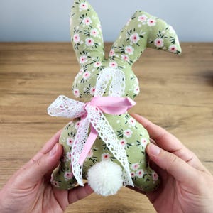 Puede incluir: Un conejito de tela verde con una cinta rosa y un borde de encaje blanco. El conejito tiene una cola de pompón blanco.