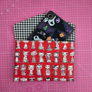 May include: A red quilted phone case with a pattern of illustrated cats in various outfits. The case has a black and white gingham lining and holds a black phone with a Halloween-themed design.
