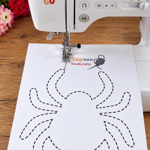 May include: A white sewing machine with a sticker reading DREAMS LET'S GO. A white sheet of paper with a black dashed spider outline and the text Etsy Narin Handicrafts is positioned under the needle, showing the start of a stitched line.