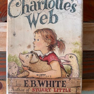 May include: A hardcover book titled Charlotte's Web by E.B. White, featuring an illustration of a young person holding a piglet, accompanied by a sheep and a goose. The cover includes the text Author of STUART LITTLE and pictures by GARTH WILLIAMS.