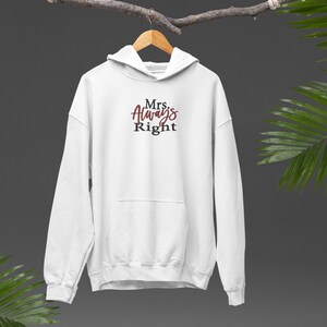 May include: A white hooded sweatshirt with the phrase "Mrs. Always Right" printed on the front in a script font. The text is in a combination of burgundy and white. The sweatshirt is hanging on a wooden hanger.
