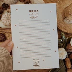 Puede incluir: Un bloc de notas rayado de color crema con las palabras "NOTES enchantées" en una fuente decorativa. El bloc de notas presenta una pequeña ilustración de oso y el texto "LA BOUILLE enchantée". También hay pequeños acentos de corazón.