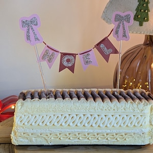 Peut inclure: Un gâteau rectangulaire avec un glaçage à la crème et une garniture marron foncé. Une bannière avec le mot "NOEL" est placée sur le dessus. Le gâteau est sur une planche de bois, avec un arrière-plan sur le thème de Noël.