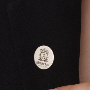 May include: A silver-colored round lapel pin with the text "RIMBERIO HOTEL HOTEL" and a crest design. The pin is affixed to a black blazer. The pin is approximately 1 inch in diameter.