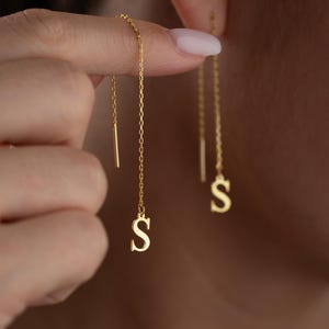 May include: Gold-colored threader earrings with the letter "S" dangling at the end of the chain. The earrings are shown in a close-up shot, highlighting the delicate design and the shiny finish of the jewelry.