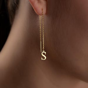 May include: A gold-colored, delicate earring with a chain and the letter "S" at the end. The earring is hanging from the ear. The background is blurred.