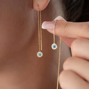 Può includere: Orecchini pendenti color oro con un piccolo ciondolo rotondo con un design a occhio blu circondato da cristalli trasparenti. Gli orecchini hanno una delicata catena, con una piccola barra all'estremità.