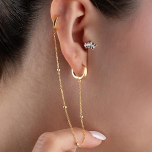 May include: A gold-toned ear cuff and hoop earring connected by a delicate chain featuring small, evenly spaced gold beads. The chain links the upper ear cuff to the lower hoop earring, creating a draped effect along the side of the ear.