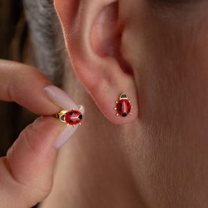 Può includere: Orecchini a bottone a forma di coccinella dorata con dettagli in smalto rosso e nero. Gli orecchini hanno la forma di coccinelle, con corpo rosso, pois neri e testa nera.