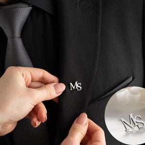 May include: A silver lapel pin with the initials "MS" is being placed on a black suit jacket. The pin is in a cursive font and is shown in a close-up shot. A gray tie is visible.