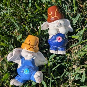 May include: Two ceramic bunny figurines, each wearing blue overalls and a brown hat. The bunnies are white with pink accents and are posed on a bed of green grass. The hats have a cracked design.