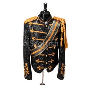 Può includere: Una giacca in stile militare con paillettes nere, caratterizzata da passamaneria dorata, bottoni metallici e una fascia diagonale. Il capo presenta spalline dorate con nappe pendenti e ricami decorativi sui polsini, esposta su un manichino.