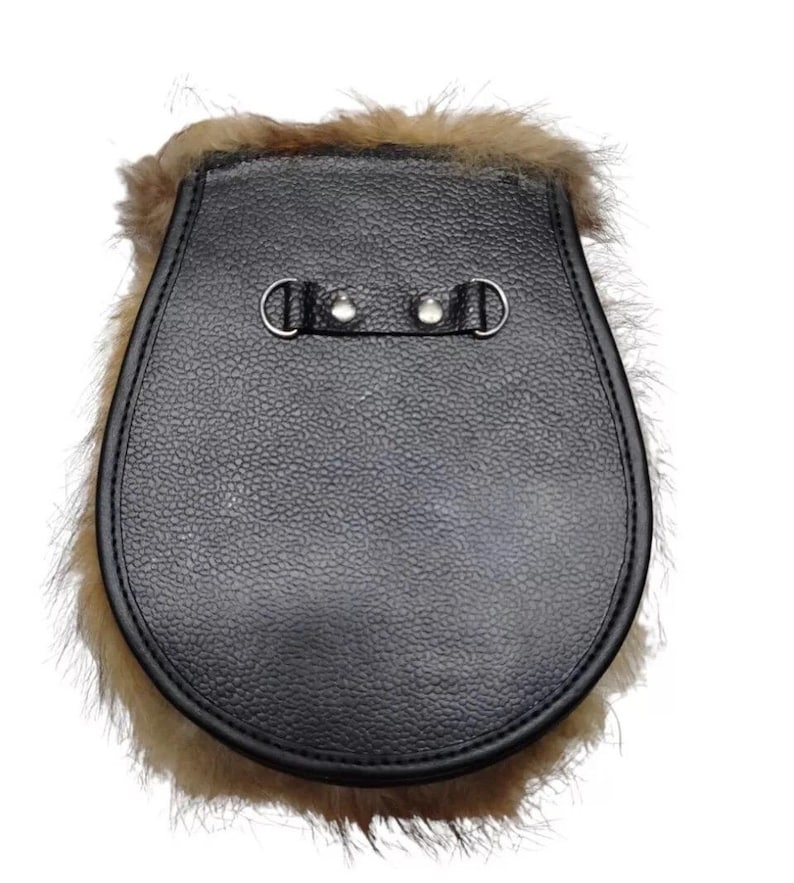 Op de afbeelding: Een zwarte leren sporran met een getextureerd oppervlak en een bontafwerking. De sporran heeft twee metalen ringen en een banddetail op de flap. Het bont is lichtbruin.
