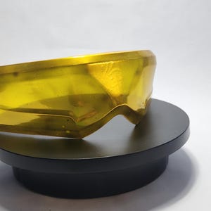 May include: A pair of yellow-tinted safety goggles, resting on a black circular stand. The goggles have a wraparound design and a smooth, reflective surface. The background is plain white.