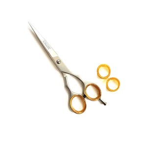 Op de afbeelding: Zilveren en goudkleurige haarschaar met twee gouden vingerringen op een witte achtergrond. De schaar heeft een gebogen handvat en een scherp lemmet. De vingerringen zijn eenvoudig en cirkelvormig.