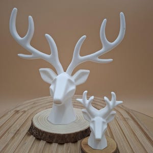 Könnte beinhalten: Zwei weiße Hirschkopfskulpturen, eine große und eine kleine, jeweils auf einer Holzscheibe montiert. Die größere Skulptur hat markante Geweihe und ist vor der kleineren positioniert. Der Hintergrund ist in einem neutralen Beigeton gehalten.