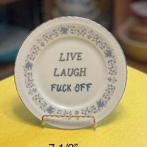 May include: A white ceramic plate with a blue floral pattern and gold trim. The plate has the text "LIVE LAUGH FUCK OFF" written in black. The plate is 7 1/2 inches in diameter and is on a gold stand.