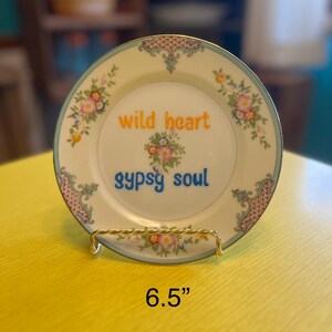 Puede incluir: Un plato decorativo con un diseño floral y las palabras "wild heart" y "gypsy soul". El plato tiene 16,5 cm de diámetro y se exhibe en un soporte de alambre dorado. El plato tiene un fondo color crema con un borde floral y de desplazamiento.