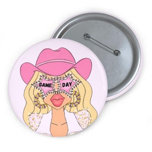 May include: A round, pink button with a cartoon illustration of a woman wearing a pink cowboy hat and star-shaped glasses. The glasses read "GAME DAY". The button has a silver pin back.