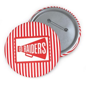 May include: A round button with a red and white striped border and background. The button features the words "GO RAIDERS" in white inside a red megaphone graphic. The back of the button is silver with a pin.