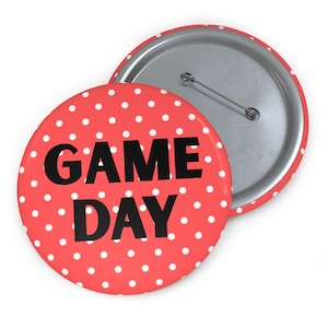 May include: A red button with white polka dots and the words "GAME DAY" in black. The button has a silver pin back and is a fun accessory for sports fans.